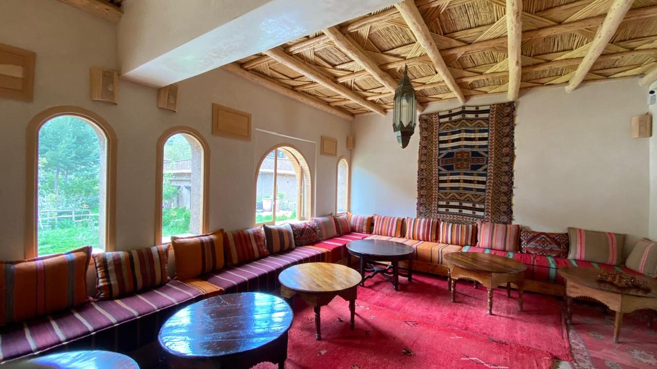Salon aux banquettes basses et coussins rayés, plafond de roseaux, tapis rouge et fenêtres en arc ouvertes sur le jardin