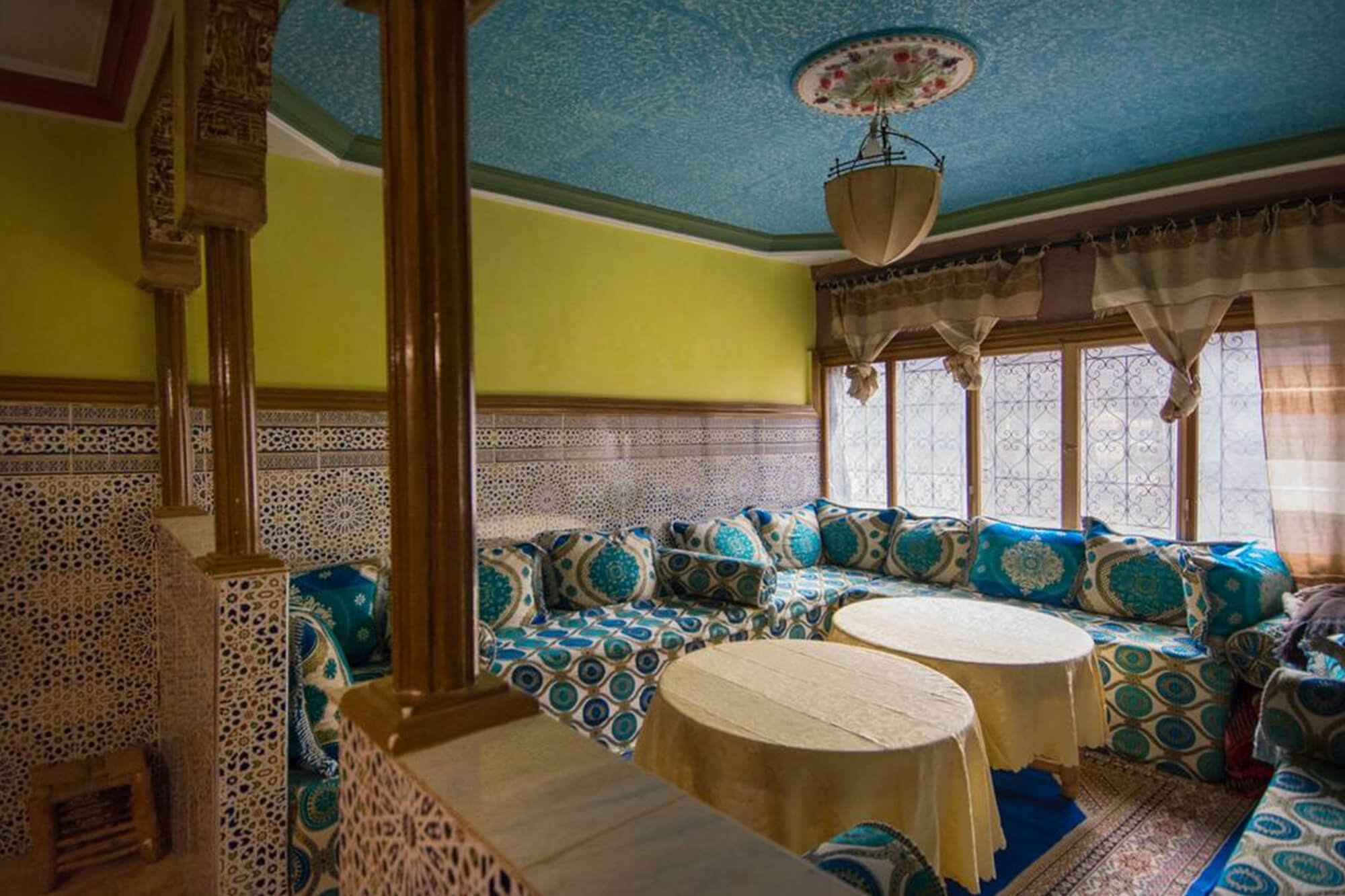 Salle à manger aux murs de zellige et plafond bleu peint, banquettes à motifs et tables rondes