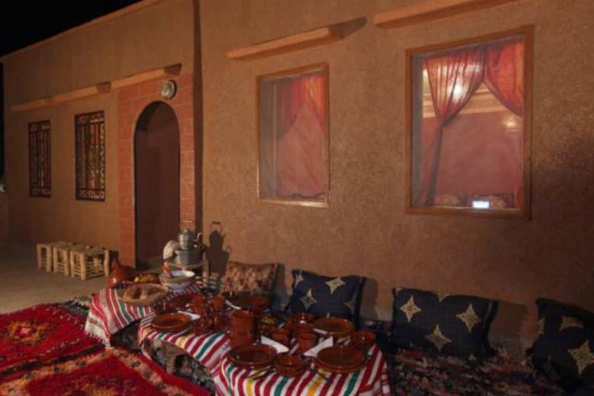 Terrasse dressée à la nuit tombée devant la façade en pisé, tapis, coussins et service à thé