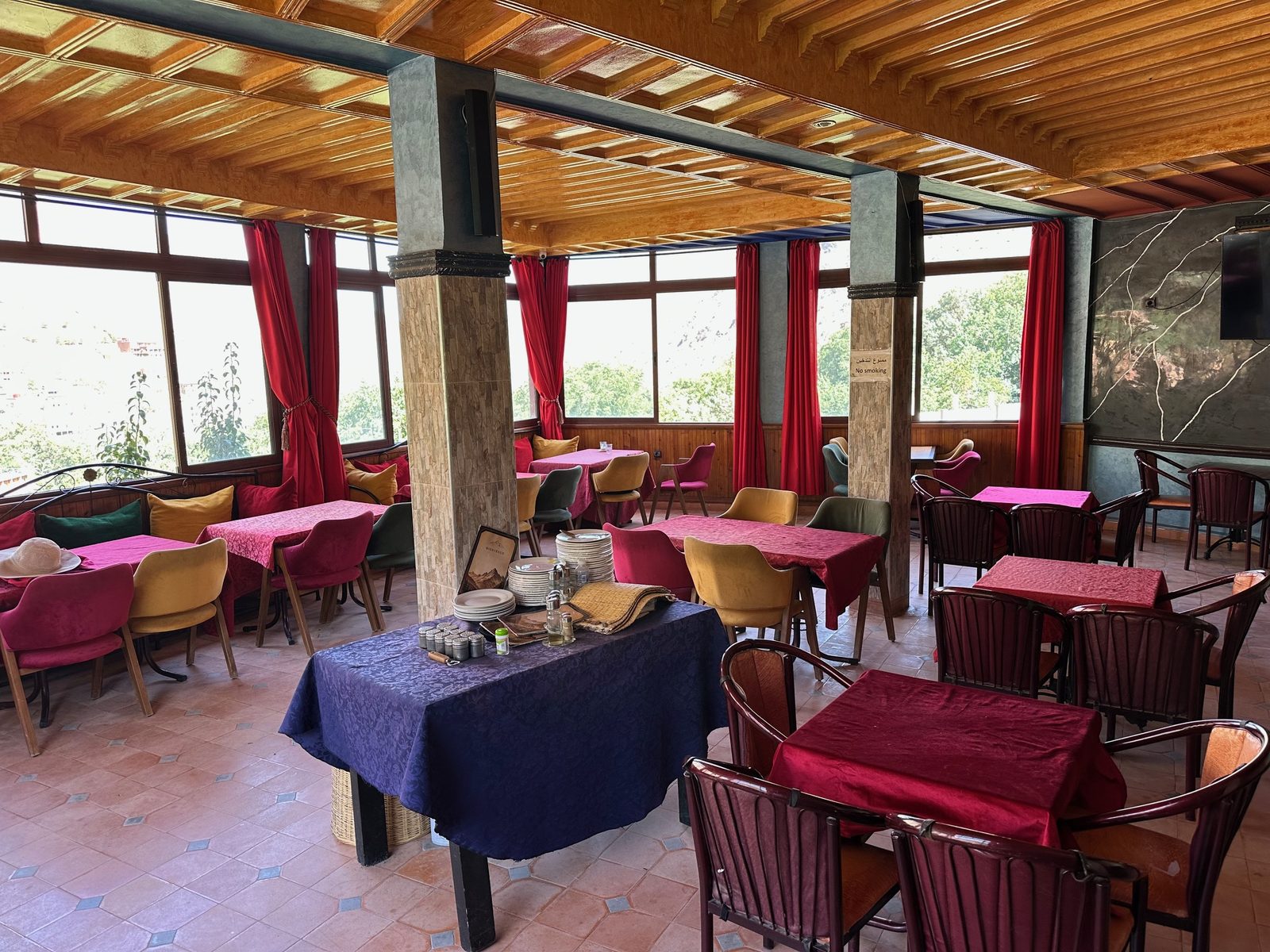 La salle vitrée du premier étage, plafond de bois clair, rideaux rouges et tables nappes de rose et de grenat, ouverte sur la vallée