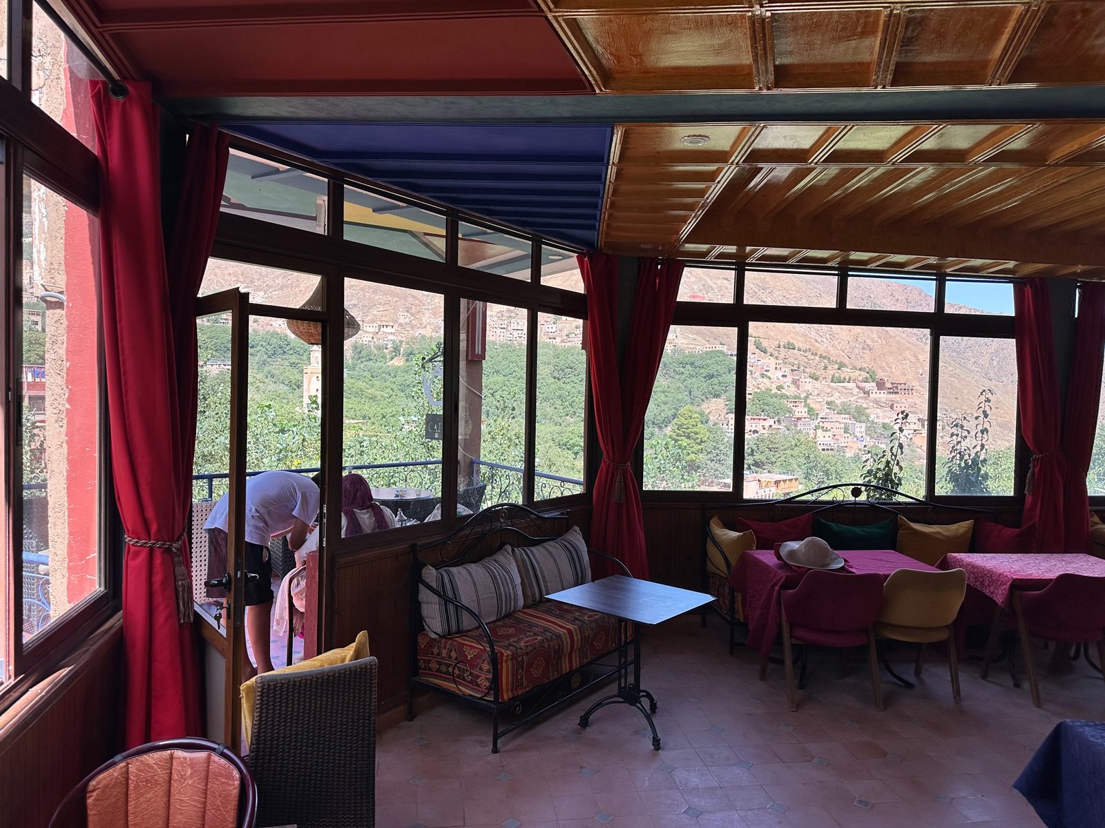Un coin salon de la salle vitrée, banquette de fer forgé et coussins rayés, avec vue sur les maisons étagées du versant