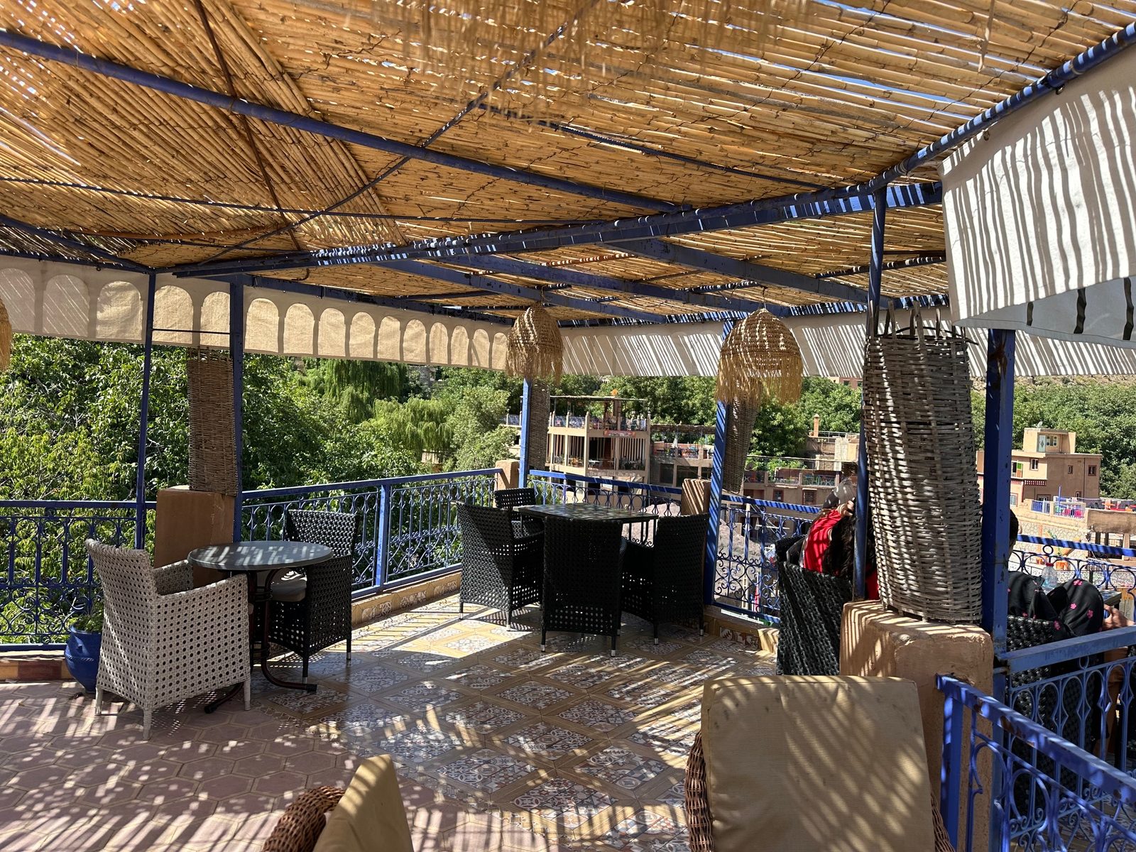 La terrasse carrelée sous la pergola bleue, suspensions d'osier et garde-corps de fer forgé au-dessus des arbres