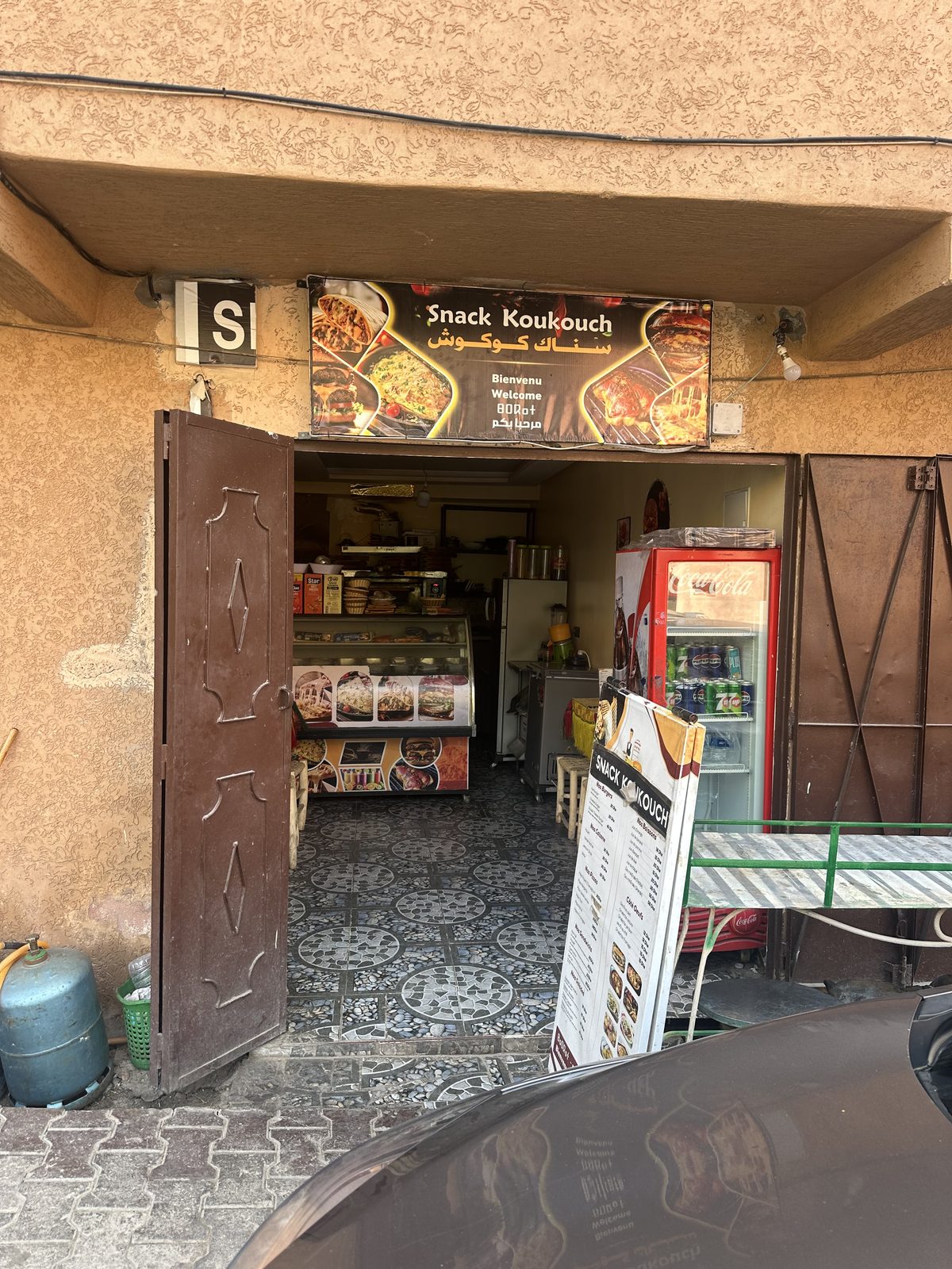 La devanture de Snack Koukouch, enseigne brune au-dessus de la porte ouverte et panneau de la carte posé sur le trottoir